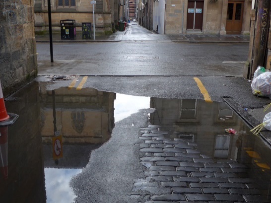 Photo looking out of the lane. The lane surface can be seen to be rough, and partially flooded. There are rubbish bags at the side of the lane.