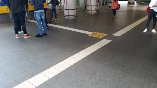 Long strips of white guideline paving on a dark grey station floor. The intention is that these can be followed along their length. There is a junction between the lines marked with a larger yellow square (which has a different texture underfoot).
