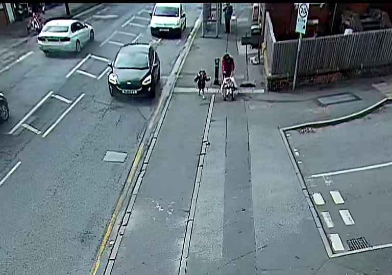 The first of four images from the fixed camera at Haddon Road. A woman is pushing a child's buggy toward the camera, with a small child walking beside her. A car is alongside them on the main road, also coming toward the camera. There is a van close behind the car on the main road.