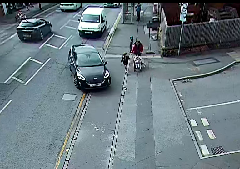 The second of four images from the fixed camera at Haddon Road. The car has begun a turn into the side road, coming closer to the pedestrians, who have continued moving forward. The woman is reaching for the child, who is out of reach.