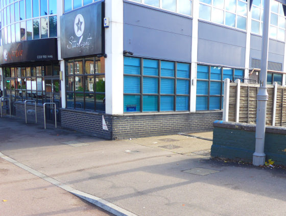 The entrance to a narrow private lane which passes alongside an industrial looking building. The footway is dropped to carriageway level at the kerb, but slopes upwards over its full width to the access lane. The footway and carriageway and access lane are all surfaced in asphalt of the same colour.
